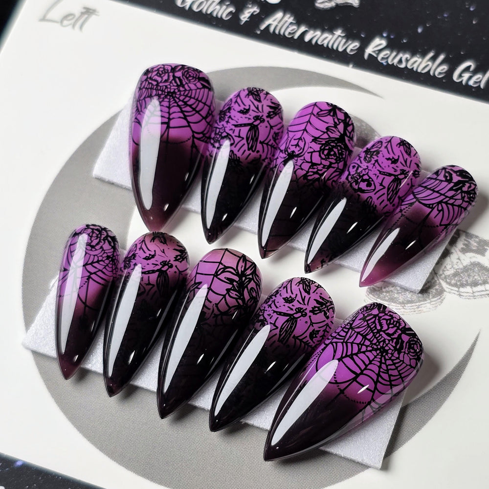 May include: Ten purple to black thermal stiletto false nails with a spiderweb design. The nails are arranged in two rows on a white surface. The text 'LD Nails Gothic & Alternative Reusable Gel' is visible in the background.