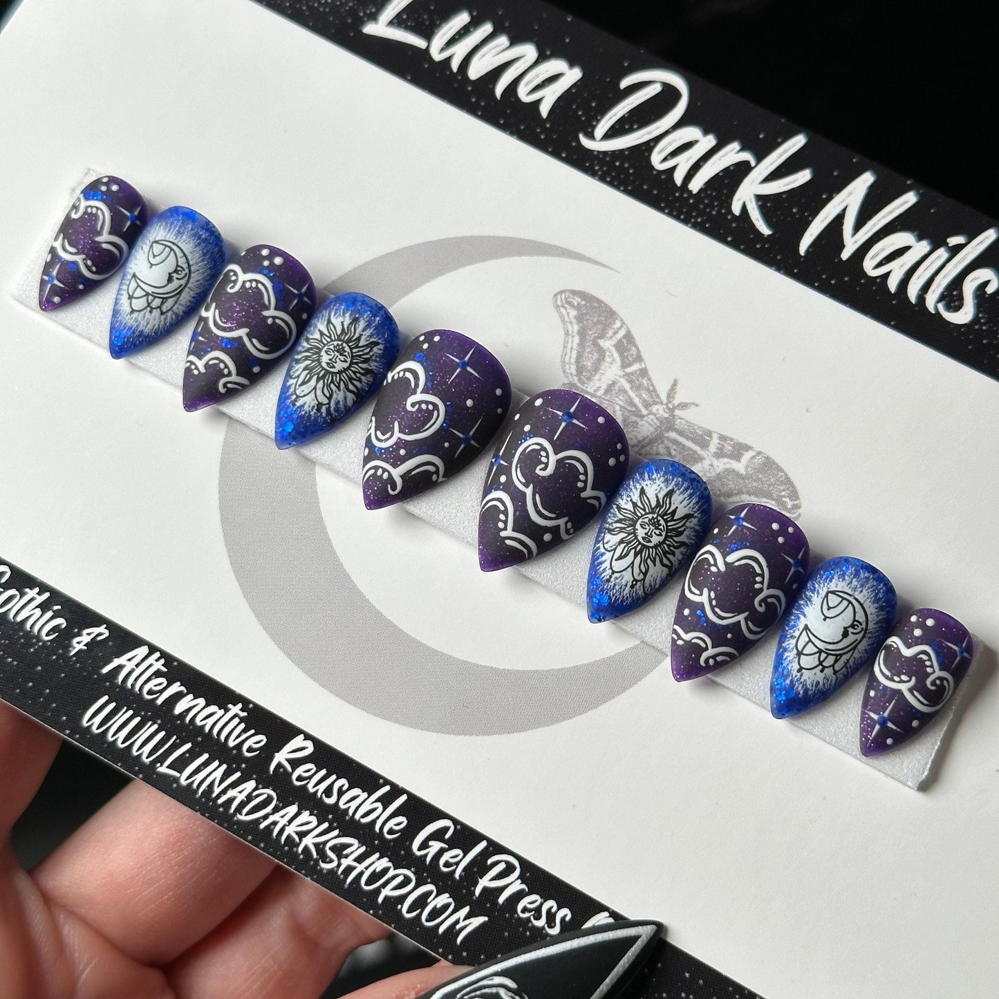 May include: Set of eight reusable gel press on nails with a dark purple base and white clouds, stars, and sun and moon designs. The nails have a blue glitter accent. The nails are displayed on a white card with the text &#39;Luna Dark Nails&#39; and &#39;Gothic & Alternative Reusable Gel Press www.lunadarkshop.com&#39;.